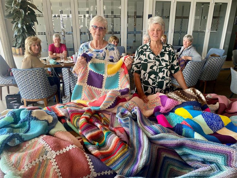 Elderly women displaying handmade knitted blankets and teddy bears for charity donation in Johannesburg