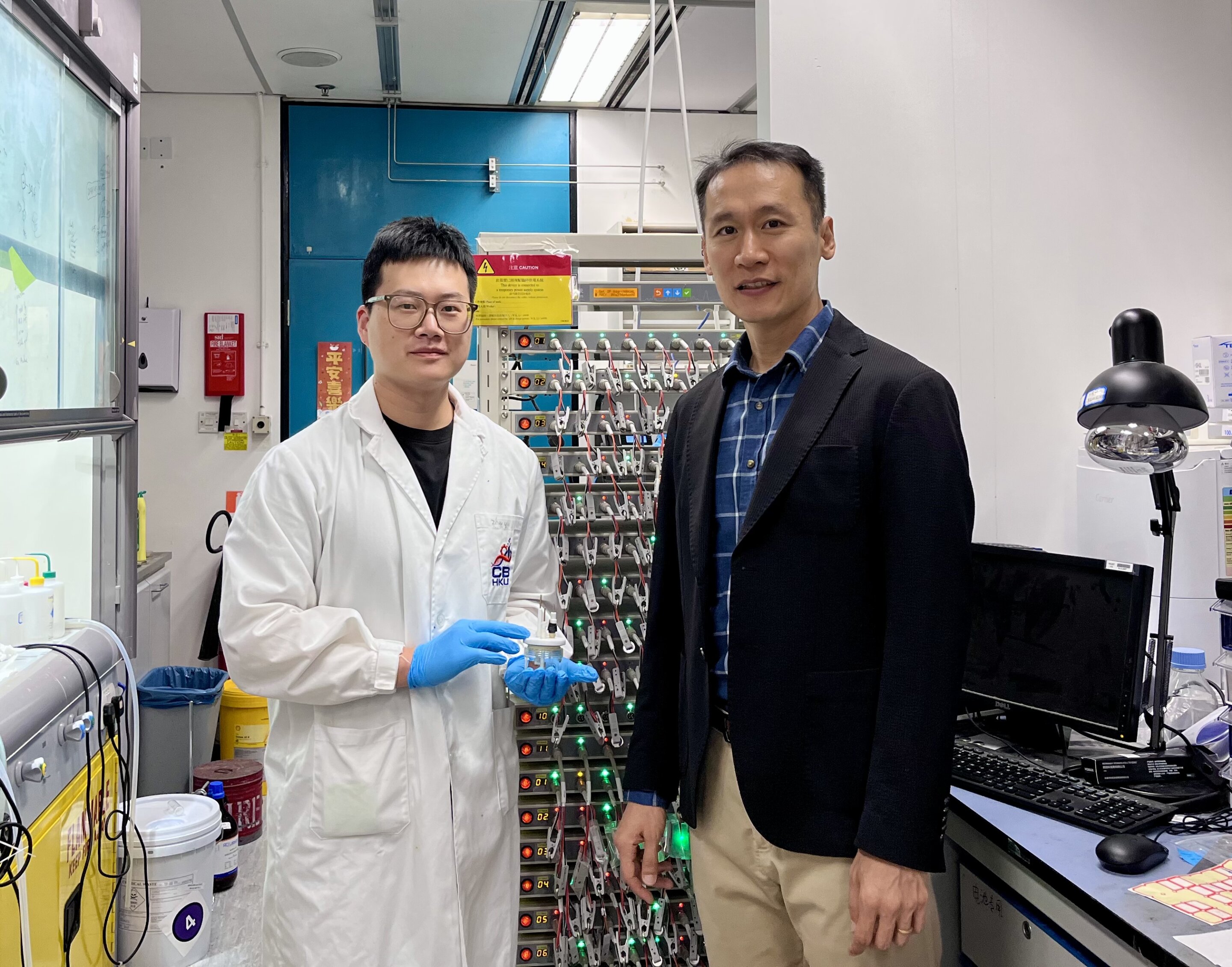 Professor Kim and student Yin holding electrochemical cell beside battery testing equipment in Hong Kong laboratory