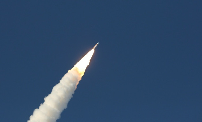 Orange and white Artemis II rocket launching into blue sky with massive flames beneath