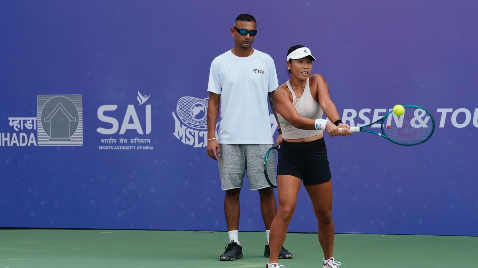 ** Indian tennis coach Mangal Sriram standing courtside watching Chinese player Fangran Tian compete in Mumbai tournament